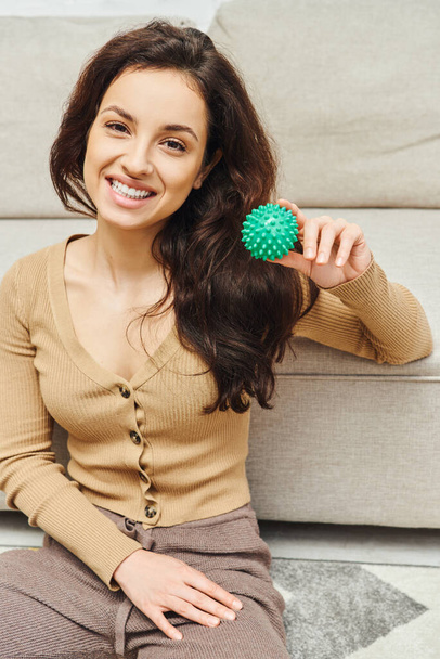 Portrait de femme brune joyeuse en vêtements décontractés souriant à la caméra et tenant une balle de massage manuelle près du canapé à la maison, massage à domicile et concept de pratiques de bien-être holistiques, équilibrant l'énergie - Photo, image