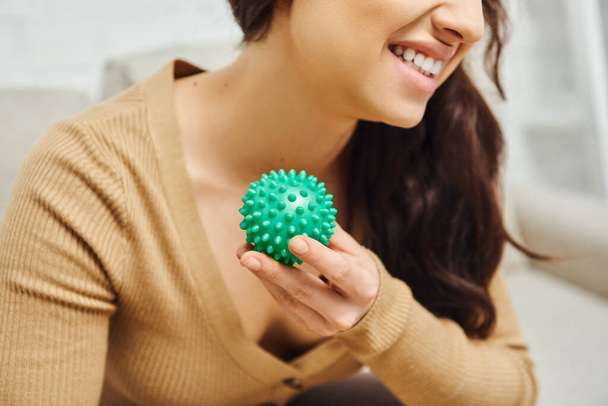 Cropped view of cheerful young brunette woman in brown jumper holding manual massage ball in blurred living room, body relaxation and holistic wellness practices, balancing energy - Photo, Image