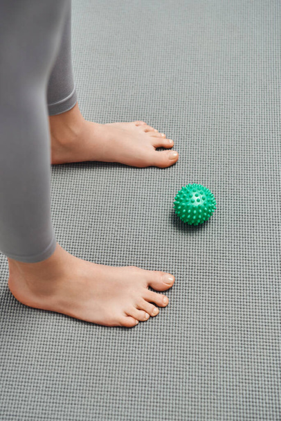 Vista superior da bola de massagem manual perto de mulher descalça em pé no tapete de fitness em casa, relaxamento corporal e práticas holísticas de bem-estar, equilibrando a energia - Foto, Imagem