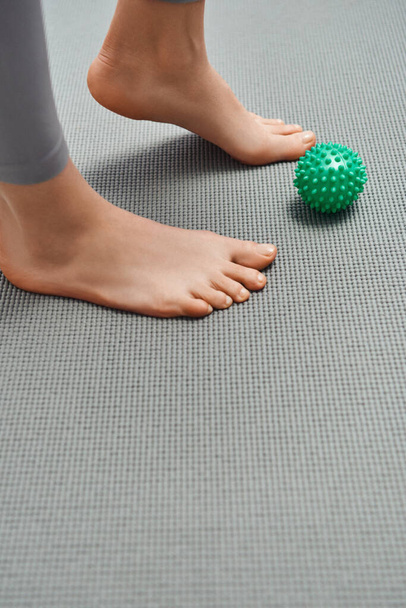 Vista ritagliata della donna scalza in piedi sul tappeto fitness vicino alla palla da massaggio manuale a casa, rilassamento del corpo e pratiche olistiche di benessere, energia riequilibrante - Foto, immagini