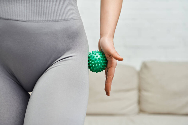 Cropped view of young woman in sportswear massaging hip with manual massage ball in blurred living room, body relaxation and holistic wellness practices, tension relief - Photo, Image