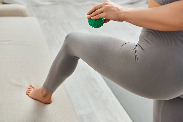 Cropped view of young woman in sportswear holding manual massage ball and standing near couch in living room at home, holistic wellness practices and body relaxation concept, tension relief - Photo, Image