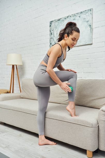 Young brunette woman in sportswear massaging leg with manual massage ball while standing near couch at home, holistic wellness practices and body relaxation concept, tension relief - Photo, Image