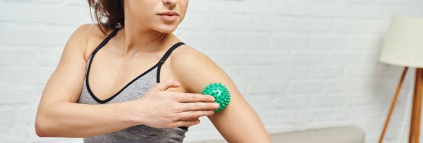Cropped view of young woman in sportswear massaging arm with manual massage ball while standing at home, balancing energy and holistic healing concept, banner, myofascial release - Photo, Image