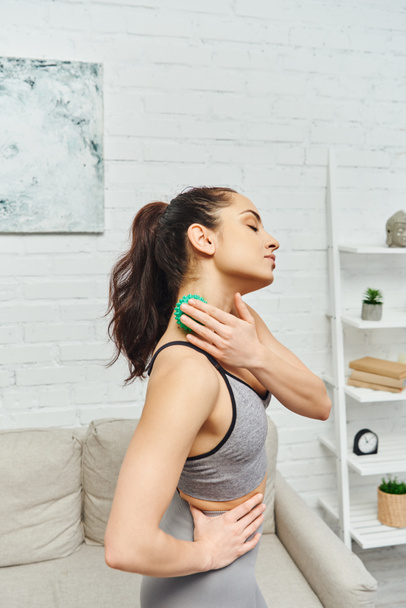 Young brunette woman in sportswear massaging neck with manual massage ball and standing in living room, balancing energy and holistic healing concept, myofascial release - Photo, Image