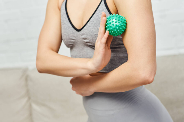 Обрізаний вид жінки в спортивному одязі масаж м'язів на руці з ручним масажним м'ячем у розмитій вітальні, балансування енергії та цілісної концепції зцілення, міофасциальний випуск
 - Фото, зображення