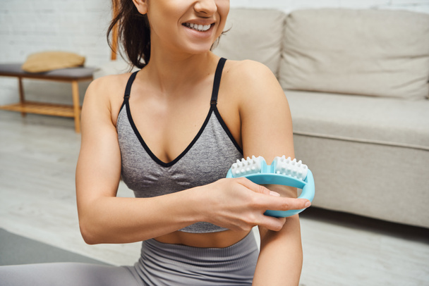 Cropped view of smiling young woman in sportswear massage arm with handle massager in floured living rom at home, balancing energy and holistic healing concept, myofascial release - Photo, image