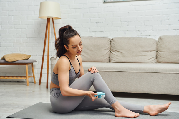 Young brunette and barefoot woman in activewear using handled massager on leg while sitting on fitness mat near couch in living room at home, inner peace and harmony concept, myofascial release - Photo, Image