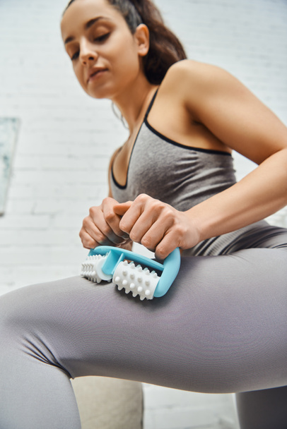 Low angle view of young blurred woman in activewear massaging leg with handled massager in living room at home, home-based massage and holistic practices concept, myofascial release - Photo, Image
