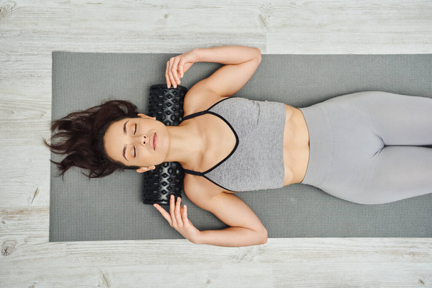Top view of young brunette woman in sportswear using roller massager on neck while lying on fitness mat at home, home-based massage and holistic practices concept, myofascial release - Photo, Image