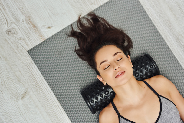 Top view of relaxed young brunette woman with closed eyes using roller massager on neck while lying on fitness mat at home, home-based massage and holistic practices concept, myofascial release - Photo, Image