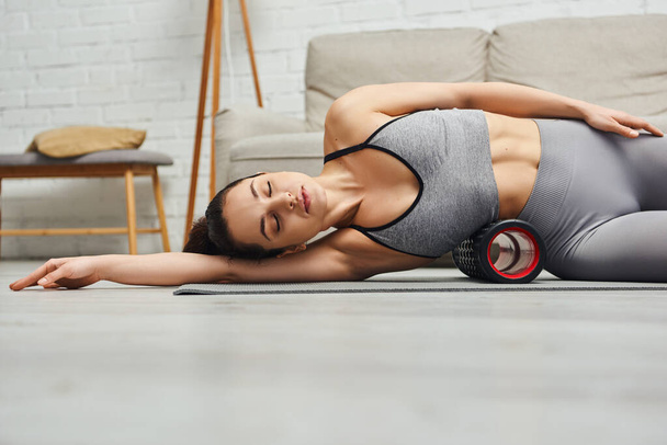 Young brunette and relaxed woman with closed eyes wearing sportswear while using roller massager on body and lying on fitness mat at home, focus on self-care and well-being concept - Photo, Image