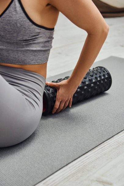 Cropped view of blurred young woman in sportswear touching modern roller massager and sitting on fitness mat at home, sense of tranquility and promote relaxation concept, myofascial release - Photo, Image