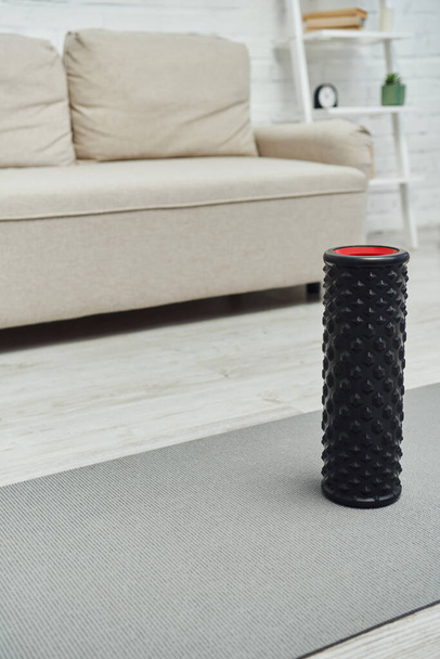 Moderner schwarzer Rollmassager steht auf Fitnessmatte neben verschwommenem Sofa im heimischen Wohnzimmer und fördert Lymphfluss und Wohlbefinden zu Hause - Foto, Bild