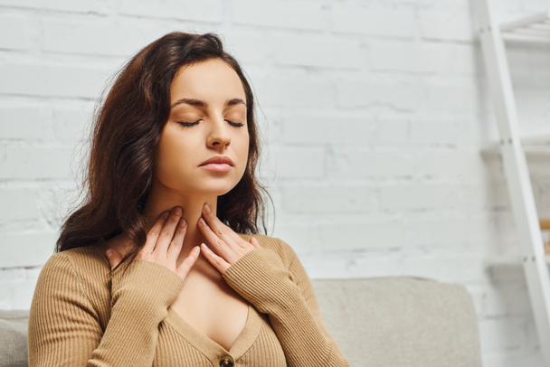 Young brunette woman in jumper checking thyroid gland on neck while sitting with closed eyes on couch in modern living room at home, focus on self-care and well-being concept, tension relief - Photo, Image