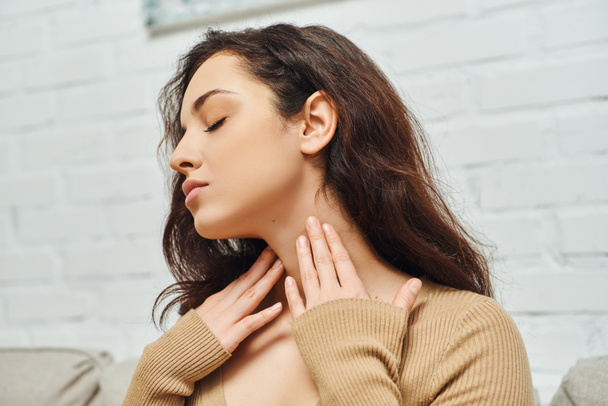 Young brunette woman in casual clothes touching neck while checking thyroid gland and sitting on couch in living room at home, focus on self-care and well-being concept, tension relief - Photo, Image