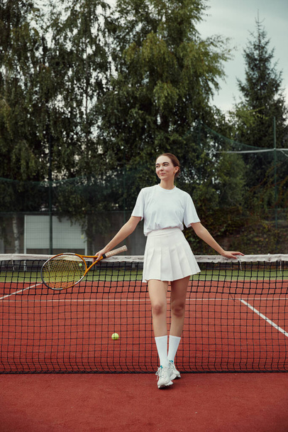 Beyaz elbiseli, tenis raketi giymiş, korttaki tenis ağında poz veren şık bir kadın. Spor Moda. - Fotoğraf, Görsel