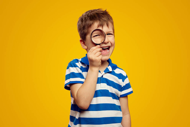 Positive curious schoolboy in casual wear looking at camera through magnifier while standing against bright yellow background - Fotografie, Obrázek
