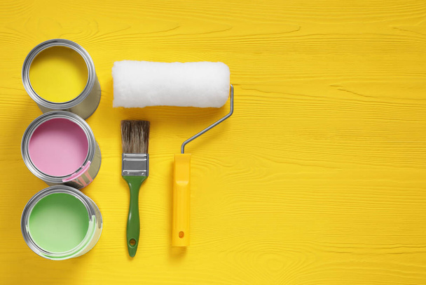 Cans of paints, brush and roller on yellow wooden table, flat lay. Space for text - Fotoğraf, Görsel