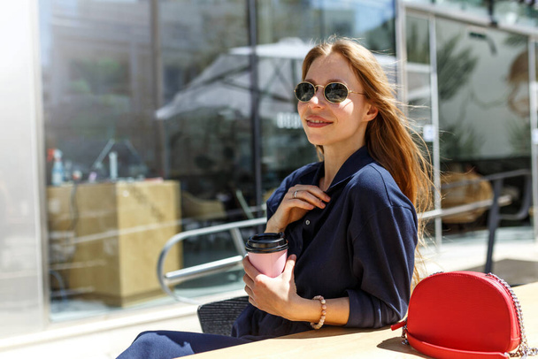 Een mooi jong hipster meisje met een kop drank zit aan een tafel van een straat zomer cafe.Take een koffierem.  - Foto, afbeelding