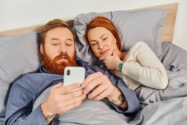 redhead couple spending time without kids, day off, husband and wife, bearded man using smartphone near woman, comfortable living, cozy bedroom, carefree, tattooed  - Photo, Image