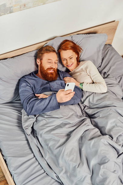 top view, happiness at home, relaxation time, tattooed couple relaxing without kids, husband and wife, bearded man using smartphone near redhead woman, cozy bedroom, carefree, screen time, day off  - Photo, Image