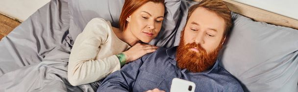 top view, networking, relaxation time, tattooed couple relaxing, weekends without kids, husband and wife, bearded man using smartphone near redhead woman, cozy bedroom, screen time, banner  - Photo, Image