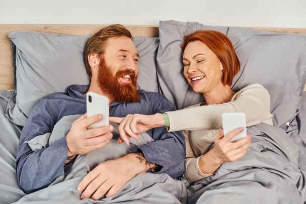 screen time, tattooed couple using smartphones, networking, relaxing on weekends without kids, husband and wife, redhead woman pointing at mobile phone of bearded man, cozy bedroom  - Φωτογραφία, εικόνα