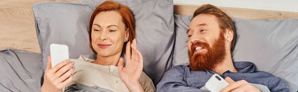screen time, tattooed couple using smartphones, relaxing on weekends without kids, husband and wife, redhead woman having video call near bearded man, cozy bedroom, waving hand, banner  - Photo, Image