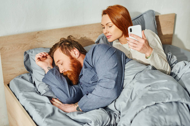 maison calme, parents seuls à la maison, femme rousse regardant son mari, homme barbu dormant près d'une femme avec smartphone, réseautage, jour de repos, réveil, tatoué, couple sans enfants  - Photo, image