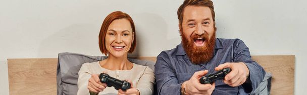 weekendy bez dzieci, rudy mąż i żona grają w gry wideo, brodaty mężczyzna i szczęśliwa kobieta trzymając joysticks, podekscytowany, zabawa w gry, małżeństwo, nowoczesny styl życia, baner  - Zdjęcie, obraz