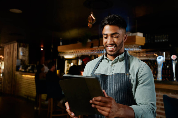 Young biracial man wearing apron smiling while using digital tablet while working as waiter in restaurant - Foto, Imagen