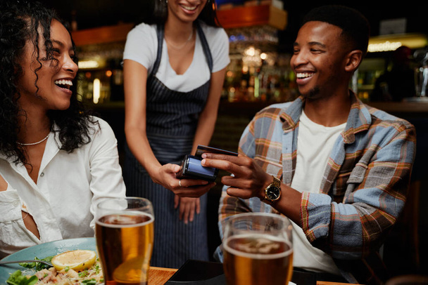 Happy multiracial friends wearing casual clothing paying waitress for dinner and drinks at restaurant - Фото, изображение