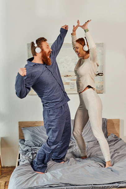 day off without kids, having fun, shared music, redhead husband and wife, happy couple in wireless headphones dancing on bed, bearded man and carefree woman, tattooed music lovers, weekends  - Photo, Image