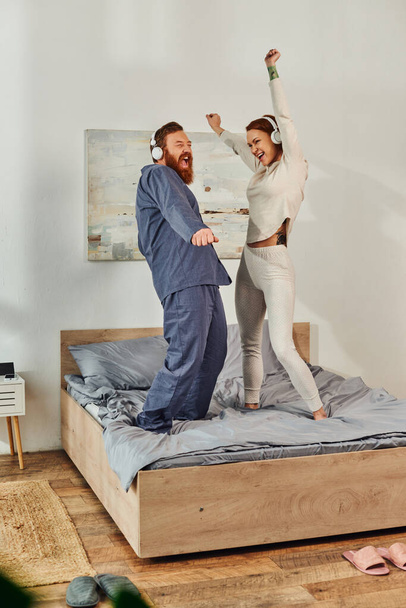 day off without kids, having fun, shared music, tattooed husband and wife, happy couple in wireless headphones dancing on bed, bearded man and carefree woman, redhead music lovers, weekends  - Photo, Image