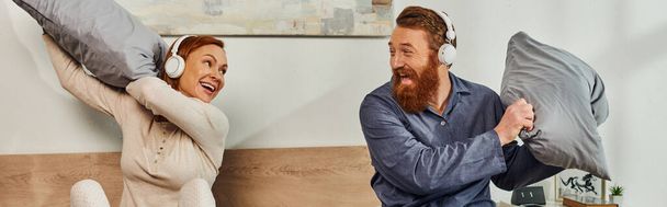 day off without kids, having fun, shared music, redhead husband and wife, happy couple in wireless headphones pillow fight, bearded man and carefree woman, tattooed music lovers, weekends, banner  - Photo, Image
