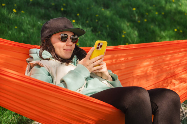 A woman in a cap lies in a hammock and looks at her phone - Foto, immagini