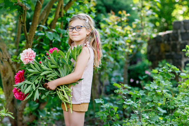 Милая очаровательная маленькая дошкольница с огромным букетом цветущих красных и розовых пионских цветов. Портрет улыбающегося дошкольника в домашнем саду в теплый весенний или летний день. Лето - Фото, изображение