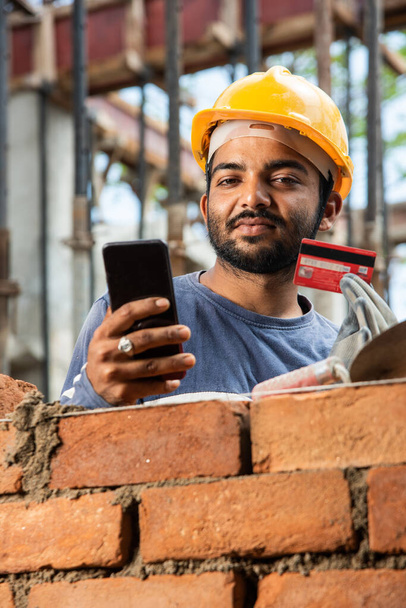 Joyeux sourire jeune travailleur indien de la construction occupé sur téléphone mobile sur le lieu de travail - Photo, image