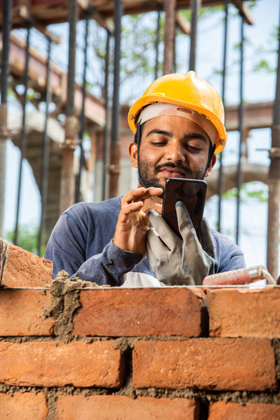 Feliz trabajo de construcción joven indio sonriente ocupado en el teléfono móvil en el lugar de trabajo - Foto, imagen