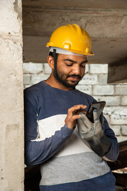 Feliz trabajo de construcción joven indio sonriente ocupado en el teléfono móvil en el lugar de trabajo - Foto, imagen
