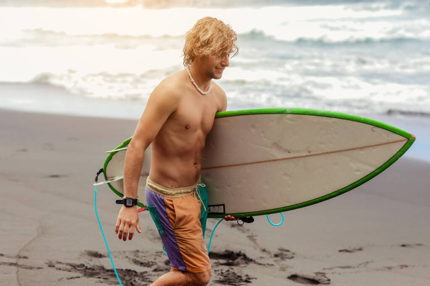 Fit młody surfer człowiek z kręconymi blond włosy z deską surfingową idzie przez ocean bawiąc się robi ekstremalne sporty wodne, surfing. Podróże i koncepcja zdrowego stylu życia. Cel podróży sportowych - Zdjęcie, obraz