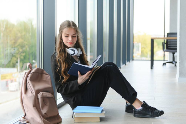 Girl smiling. Beautiful schoolgirl smiling while sitting near window and reading book. - Zdjęcie, obraz