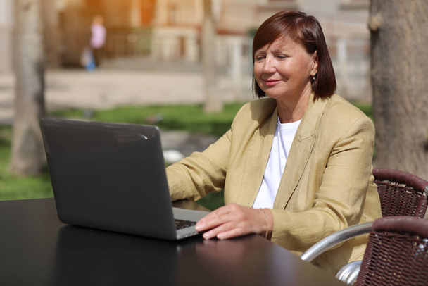 Elegante volwassen zakenvrouw werkt en typt op laptop, het maken van een oproep op de smartphone in een outdoor cafe in de stad. Concept van communicatie op afstand vanuit de openbare ruimte, digitale freelance, moderne levensstijl - Foto, afbeelding