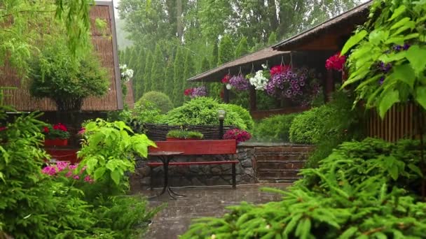 Lluvia en un pintoresco jardín con flores, un banco y una mesa en verano - Metraje, vídeo