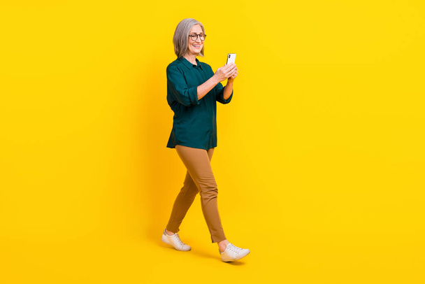 Pełnowymiarowy portret pozytywnej pani używać smartfona spacery izolowane na żółtym tle koloru. - Zdjęcie, obraz