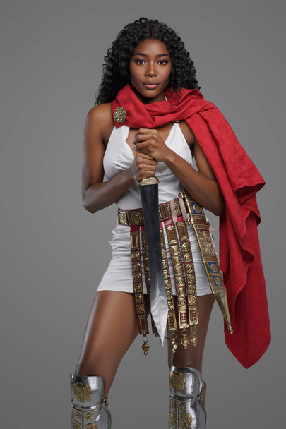 Prachtig afro-Amerikaans model in witte tuniek met een prachtige rode cape versierde riemgraven terwijl ze vol vertrouwen poseren met gladius zwaard tegen grijze achtergrond - Foto, afbeelding