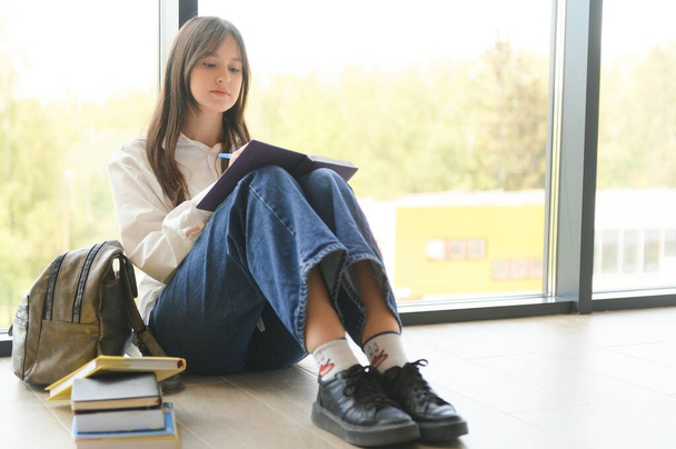 Girl smiling. Beautiful schoolgirl smiling while sitting near window and reading book. - Foto, Imagem