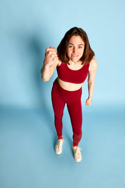 Top view full-length portrait of young emotional girl showing fist, expressing anger and annoyance against blue studio background. Irritation. Concept of youth, human emotions, lifestyle, ad - Fotografie, Obrázek
