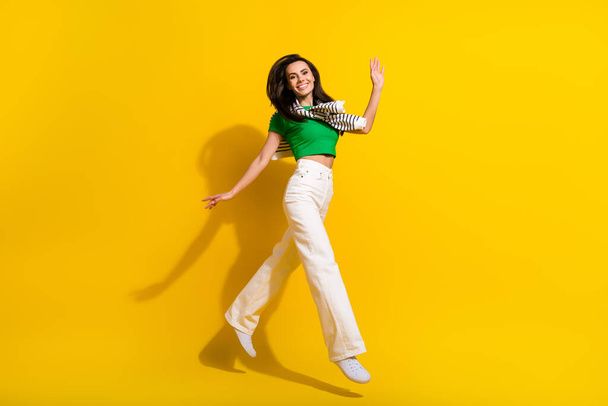 Full length photo of excited friendly lady wear white trousers jumping high walking waving arm isolated yellow color background. - Fotó, kép